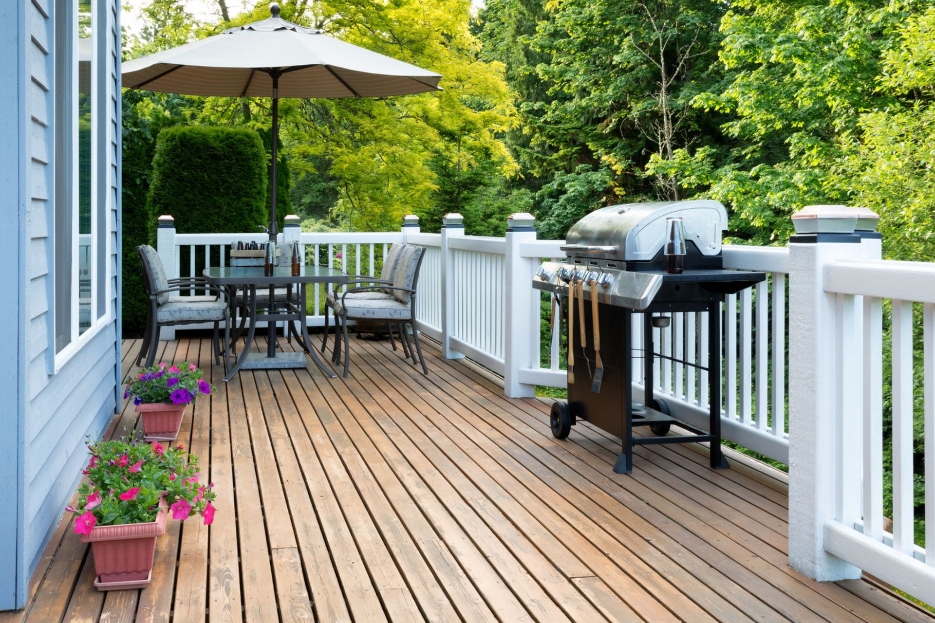 Wooden deck with white railing, patio table and grill, surrounded by green trees and potted flowers.