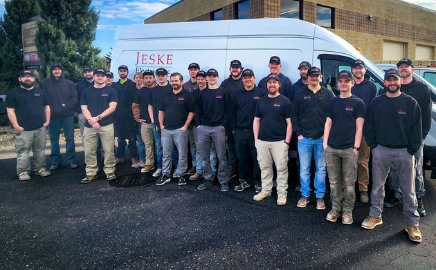Group of people in black shirts posing in front of a white van and industrial building outdoors