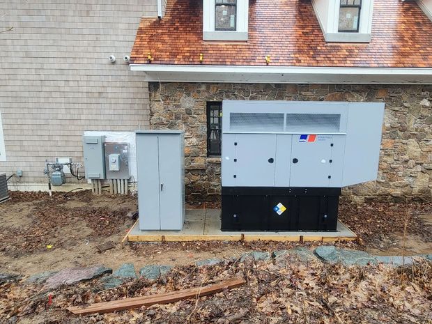 Outdoor generator and utility boxes beside a brick building, with dirt ground and leaf debris in front.