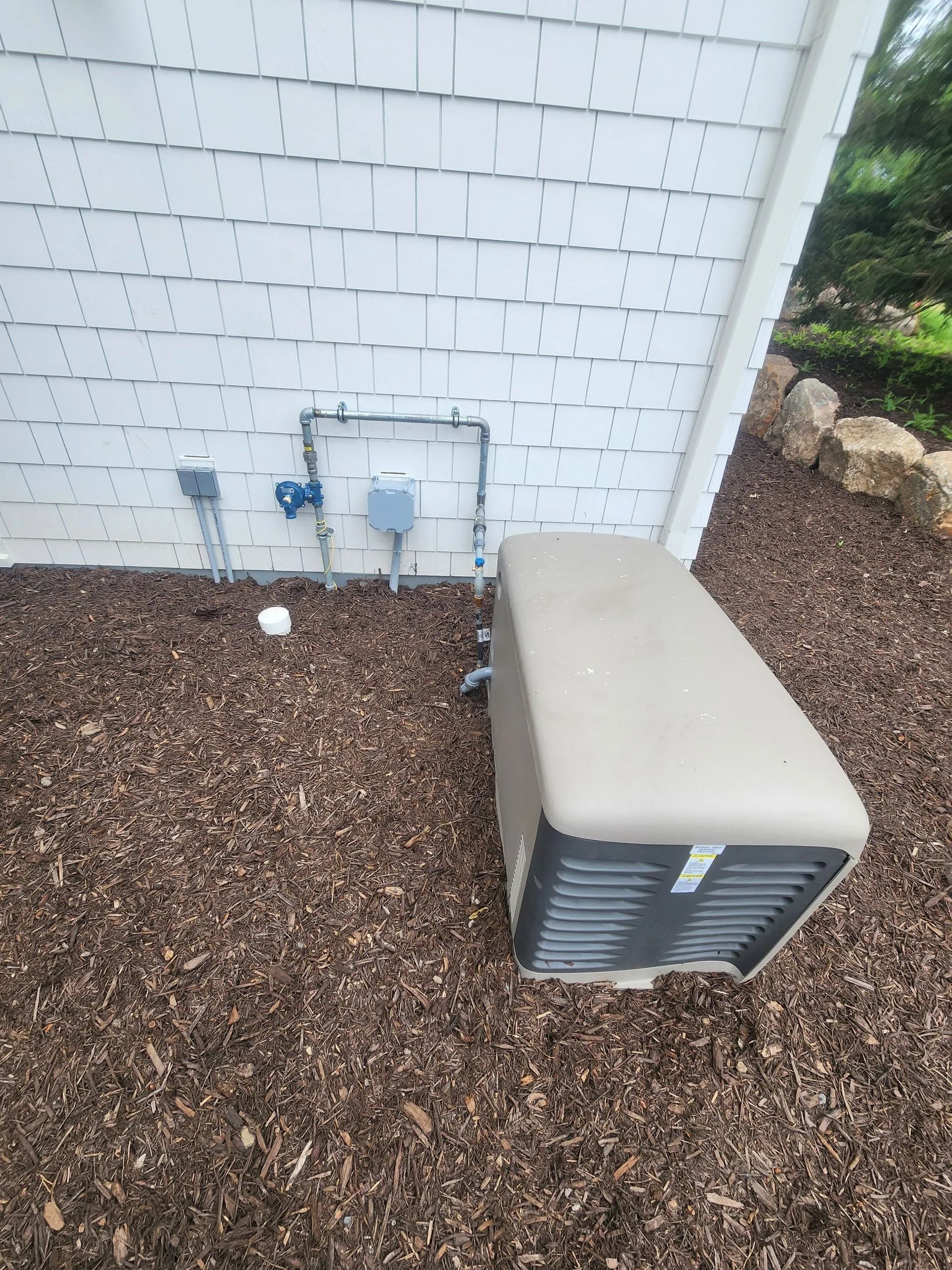 Outdoor HVAC unit beside a white house wall on a bed of gravel, with utility boxes nearby