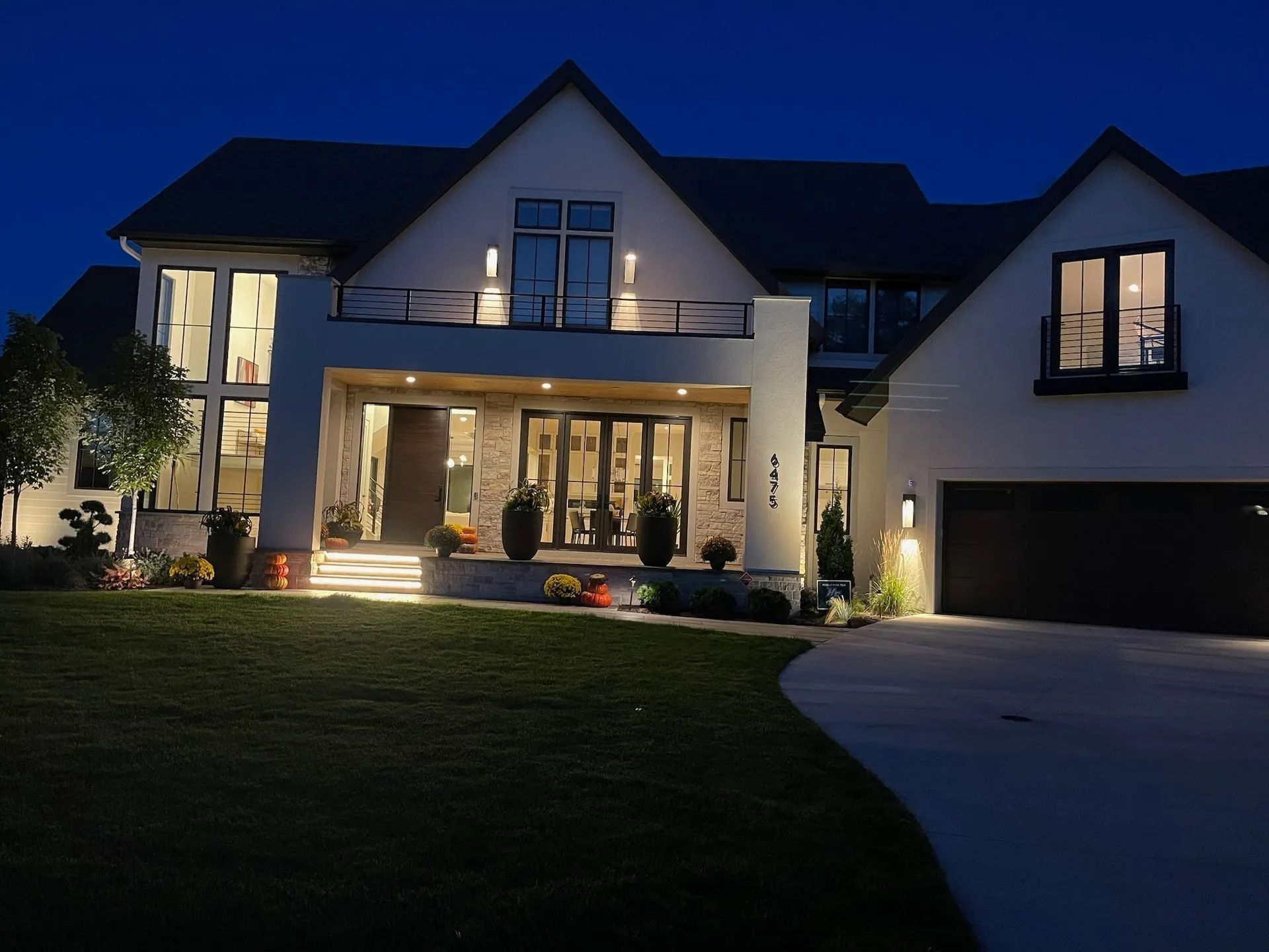 Large modern house at dusk with warm exterior lights and a dark front lawn and driveway