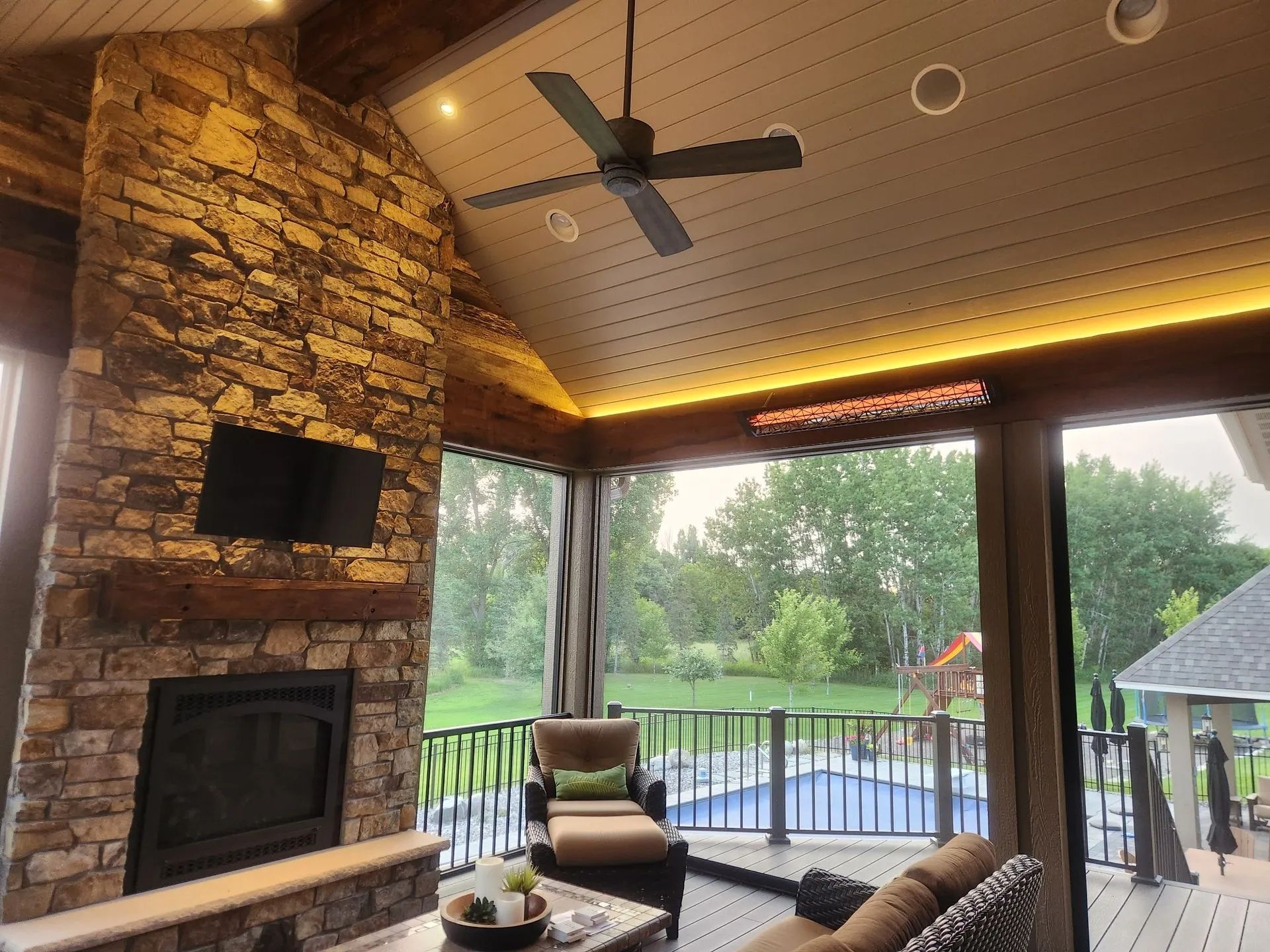 Covered patio with stone fireplace, ceiling fan, TV, and a view of the backyard pool and trees.