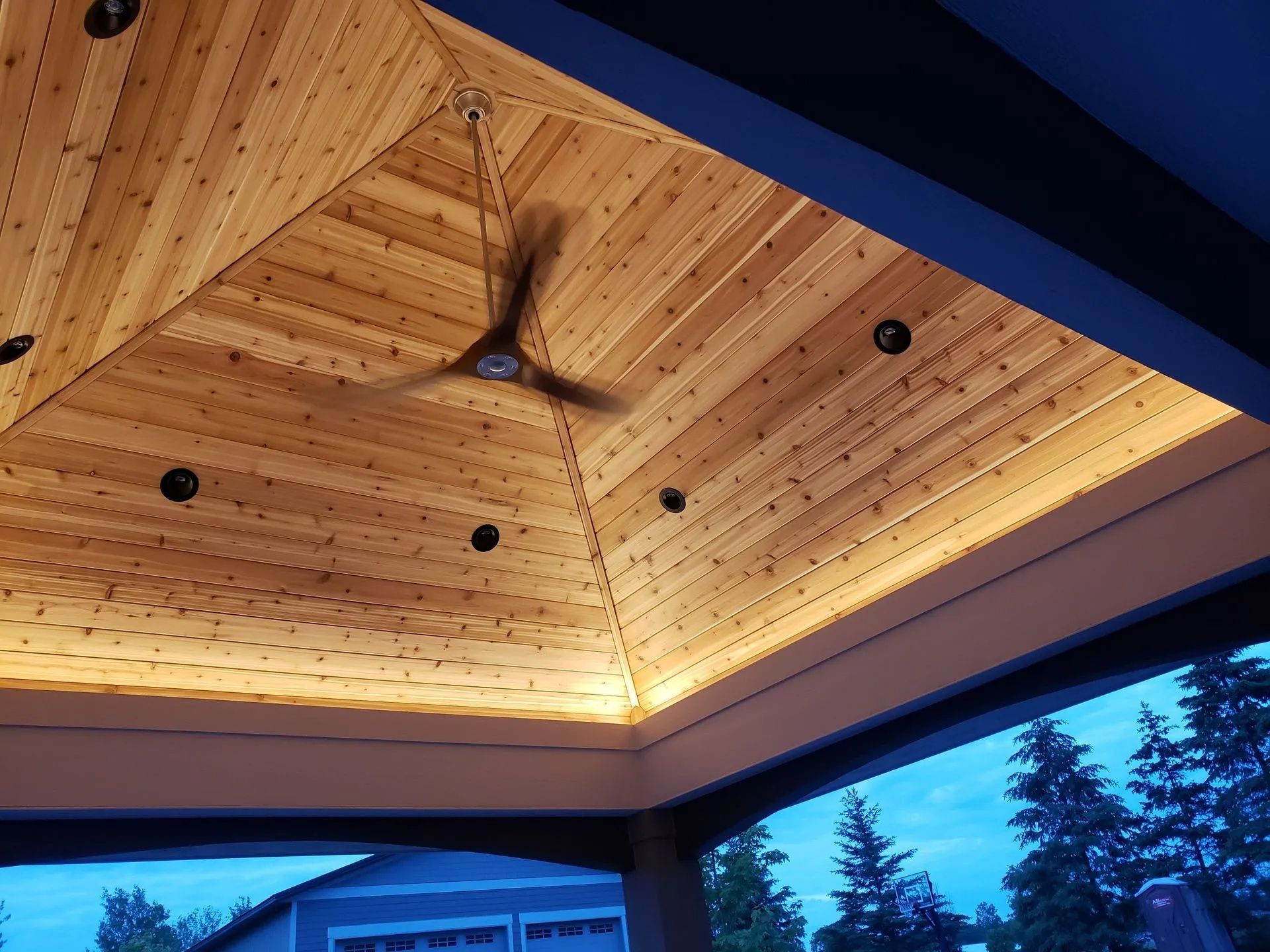 Wood-paneled ceiling with recessed lights and a fan above a dark outdoor deck at dusk