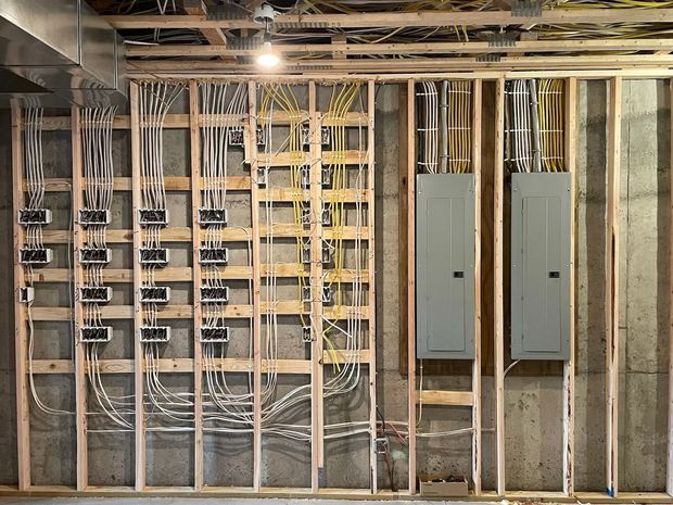 Unfinished basement wall with exposed wiring, studs, and two gray electrical panels.