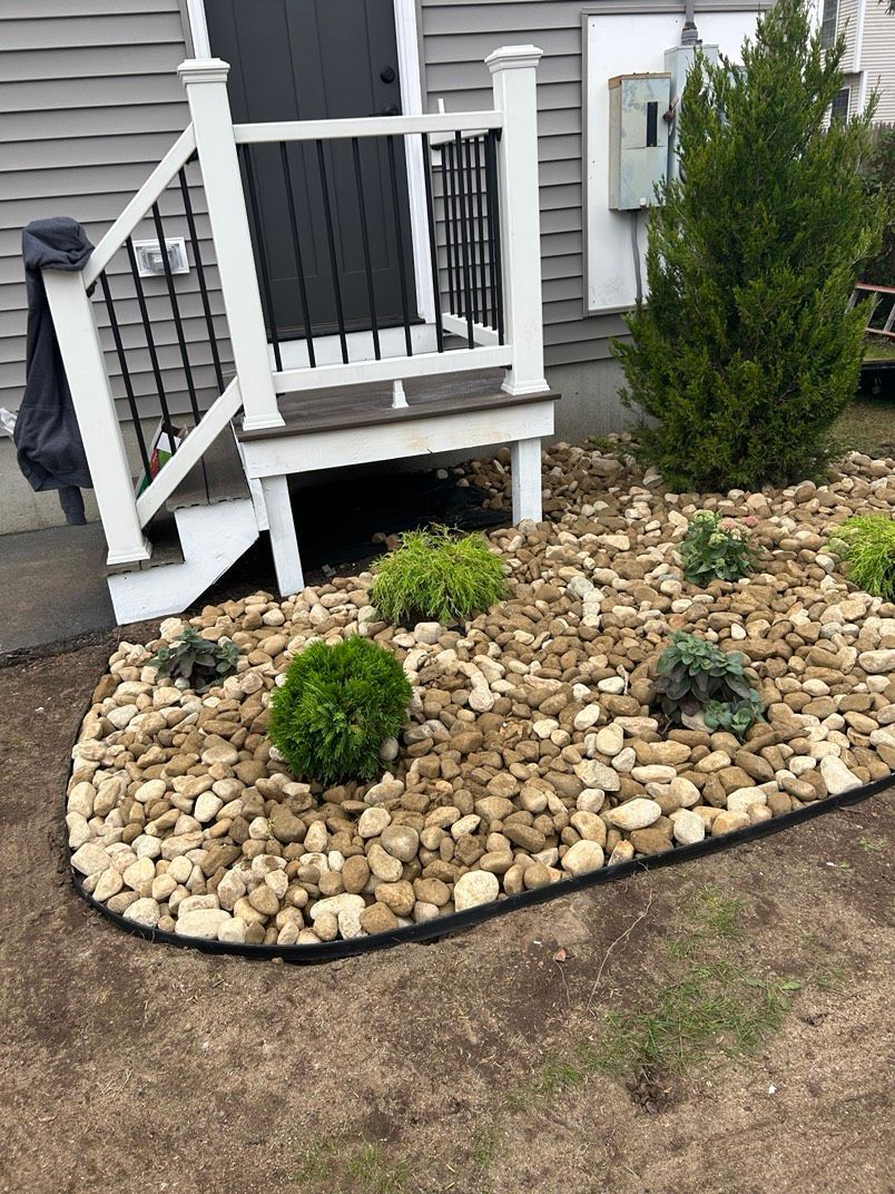 Small front yard landscaping with new plants. A short deck with white railing. Gray siding and dark door.