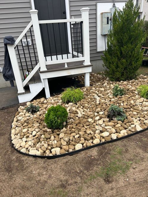 Small front yard landscaping with new plants. A short deck with white railing. Gray siding and dark door.