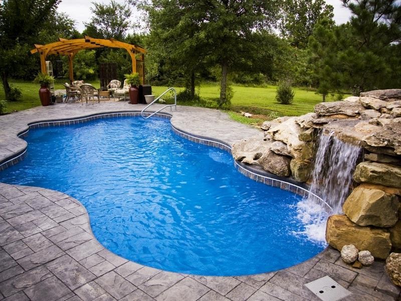 Swimming pool with waterfall, surrounded by stone patio, lawn, and pergola with seating.