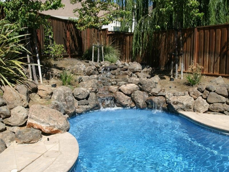 Pool with a rock waterfall feature in a backyard with a wooden fence.