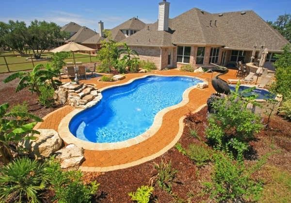 Backyard pool with a house in the background; Blue water, brown landscaping and rocks.