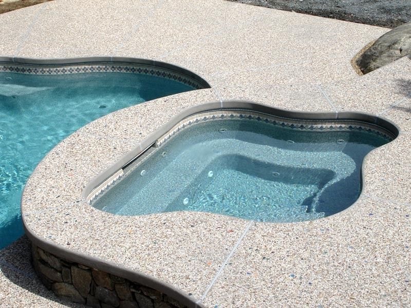 Pool and spa with light-colored speckled concrete decking. Blue water in pool and spa, steps in spa.