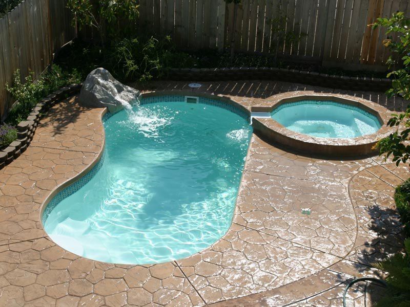 Small, kidney-shaped swimming pool with connected hot tub, waterfall feature, and textured concrete patio.