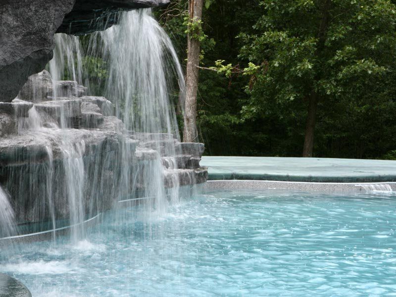 Waterfall cascading into a turquoise swimming pool, surrounded by stone and trees.