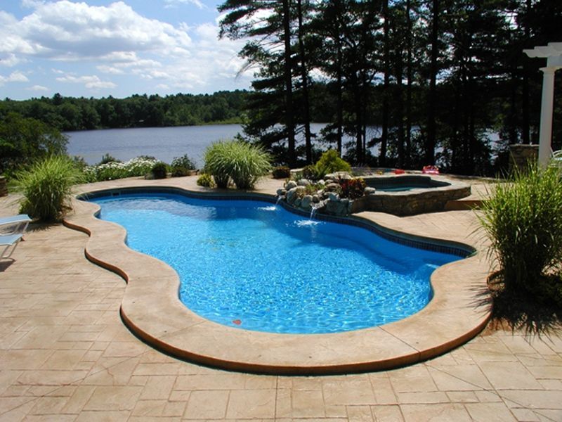 Pool overlooking a lake, with waterfall feature and built-in hot tub. Blue water, concrete patio.