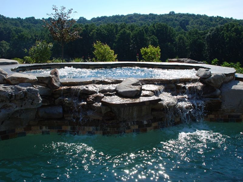 Hot tub with cascading waterfall flowing into a pool, with trees in the background under a blue sky.