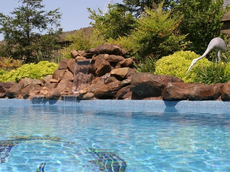 Swimming pool with waterfall feature and a statue of a heron in a landscaped garden.