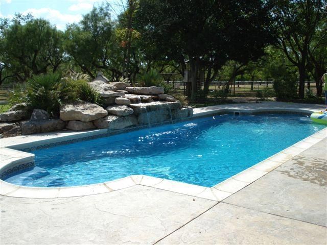 Swimming pool with waterfall feature, surrounded by concrete patio and greenery.