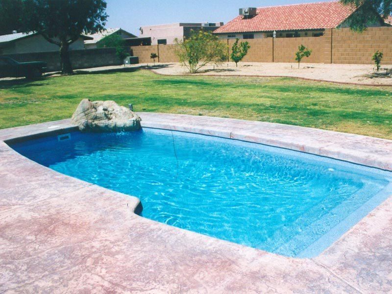 Rectangular blue pool with rock waterfall, surrounded by concrete patio and green lawn. Houses in background.