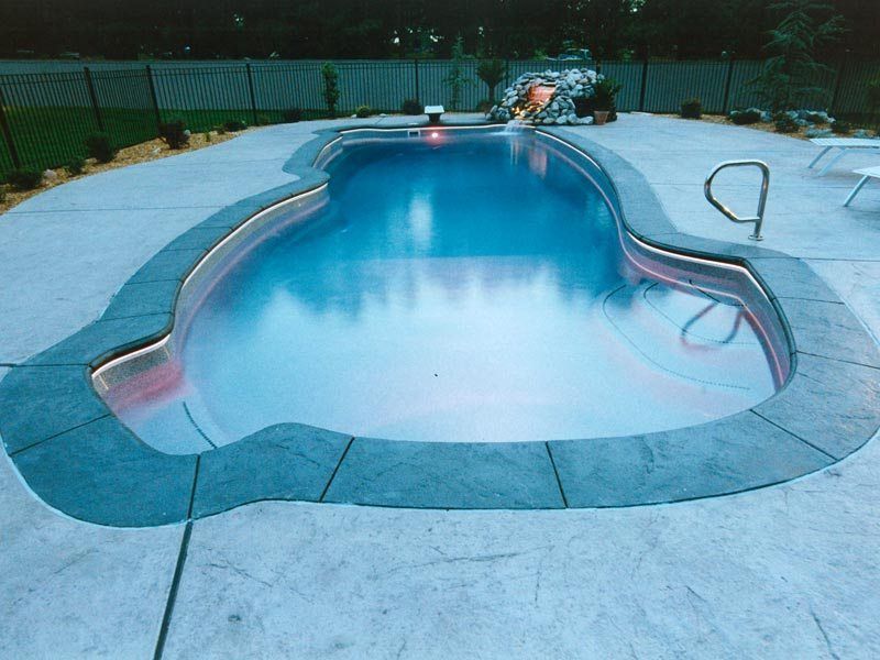 Swimming pool with a curved shape, surrounded by a concrete deck. Back wall has a small waterfall feature.