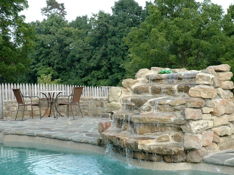 Pool with rock waterfall, patio with table and chairs, trees in background.