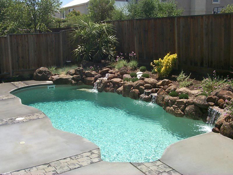 Pool with turquoise water and rock waterfall feature, set in a backyard with wooden fence.