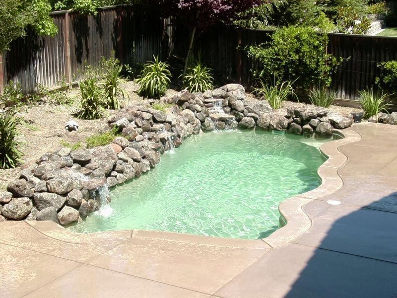 Small backyard pool with a rock waterfall feature, surrounded by concrete and landscaping.