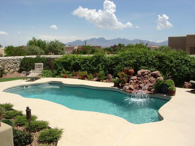 Swimming pool with waterfall feature, surrounded by landscaping, under a blue sky with mountains in the background.
