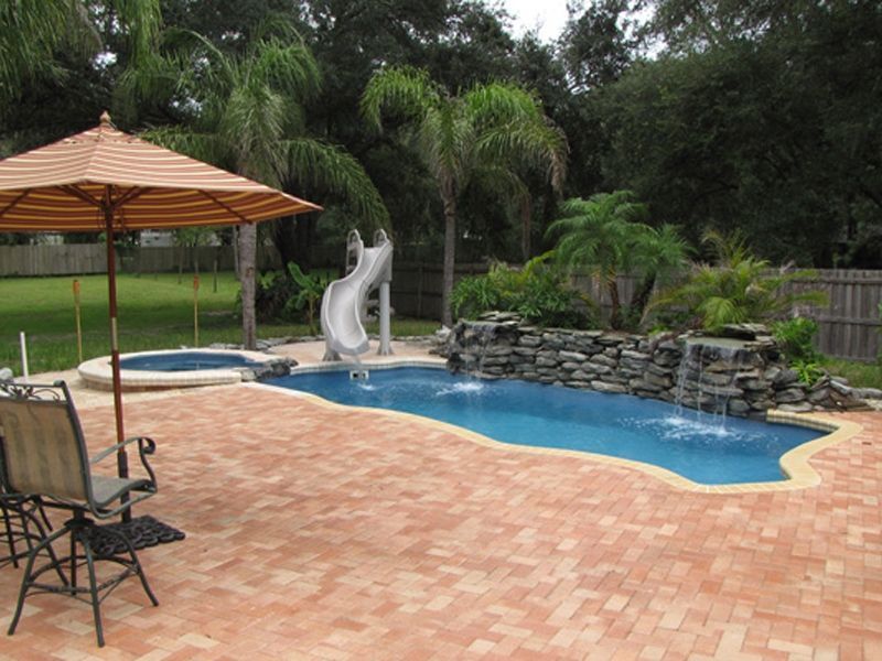 Backyard with a swimming pool, spa, brick patio, umbrella, slide, and waterfall feature.