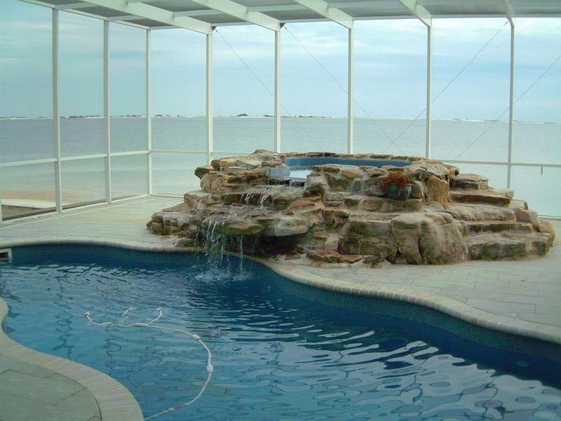 Swimming pool with waterfall feature, enclosed by white-framed glass walls.
