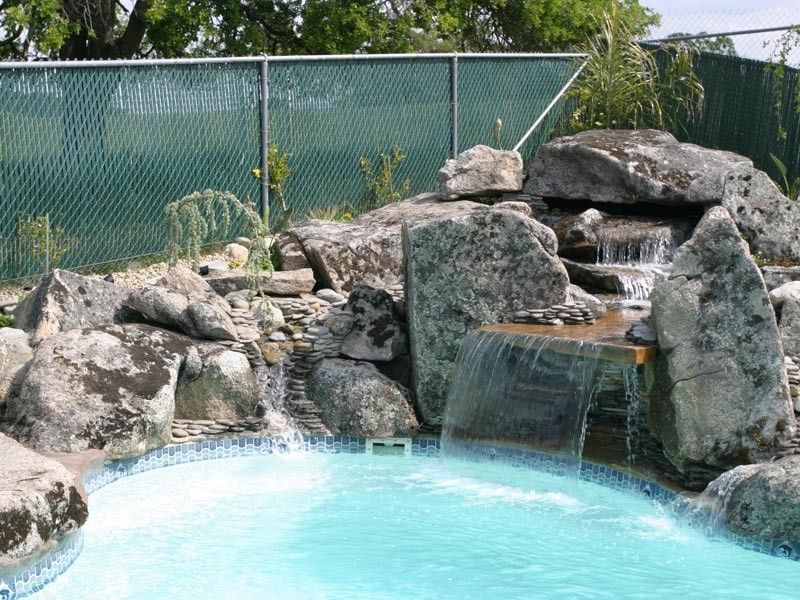 Pool with waterfall cascading over rocks; green chain-link fence in background.
