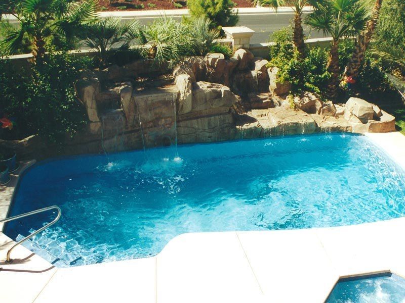 Blue pool with waterfall feature, white deck, and greenery in a sunny setting.