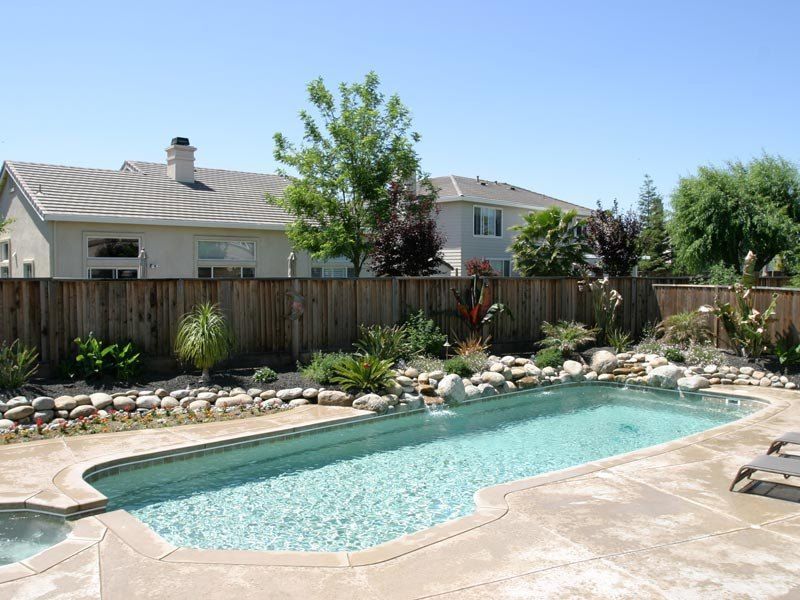 Backyard pool with light blue water surrounded by concrete, rocks, and plants. Houses in background.