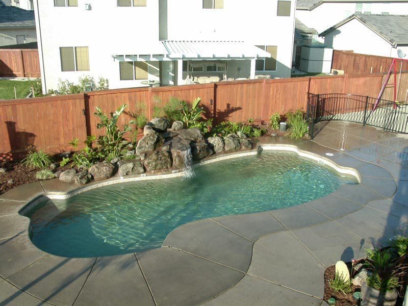Backyard pool with waterfall feature, surrounded by concrete, landscaping, and a wooden fence.