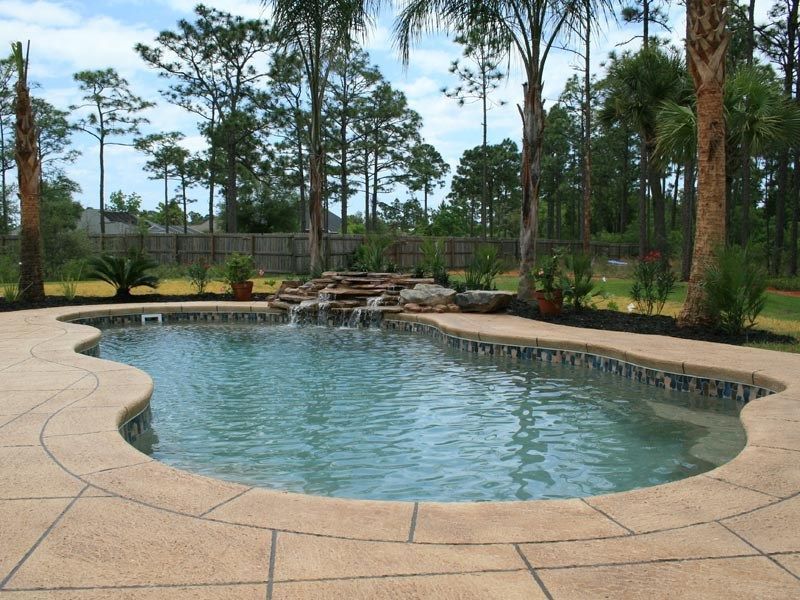 Swimming pool with waterfall feature, surrounded by trees and a paved patio.