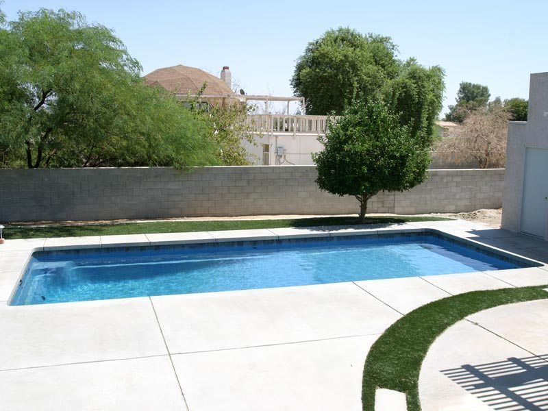 Rectangular pool in a backyard with concrete patio, grass, trees, and a concrete wall.
