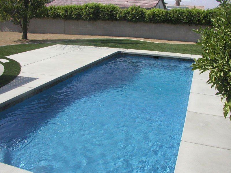 Rectangular blue swimming pool surrounded by concrete and lawn, in a backyard setting.