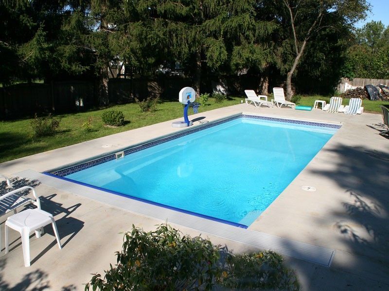 Rectangular swimming pool surrounded by concrete, basketball hoop, lawn chairs. Blue water, sunny outdoor setting.