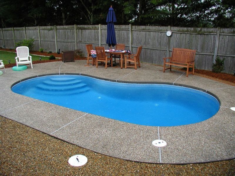 Oval-shaped swimming pool in backyard with patio furniture and umbrella, surrounded by gravel and a wooden fence.