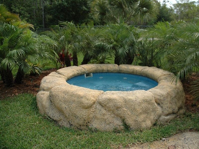 Circular hot tub surrounded by large rockwork, set in a grassy yard with palm trees.