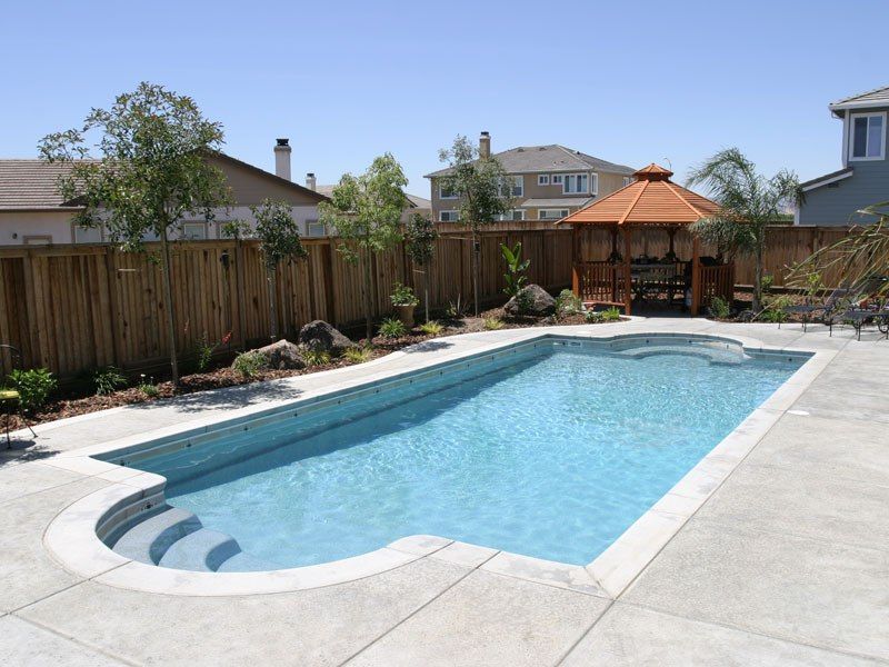 Rectangular swimming pool with curved steps and adjacent gazebo, concrete patio, wooden fence, and houses in the background.