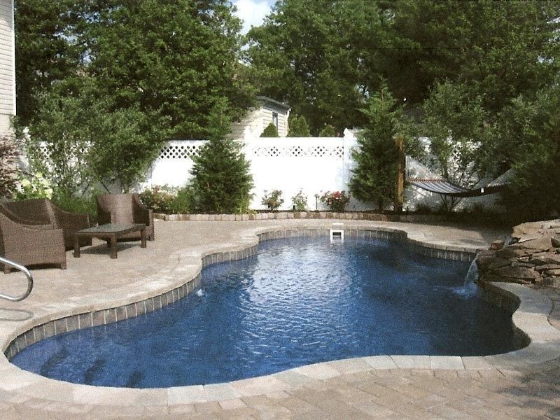 Backyard pool with stone patio and seating, surrounded by trees and a white fence.
