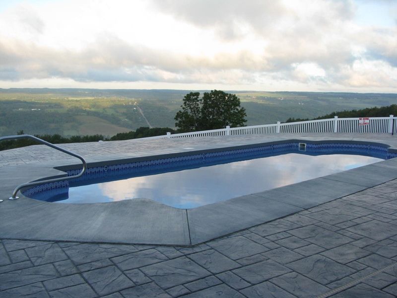 Rectangular swimming pool overlooking a valley, blue water, gray deck, white fence, cloudy sky.