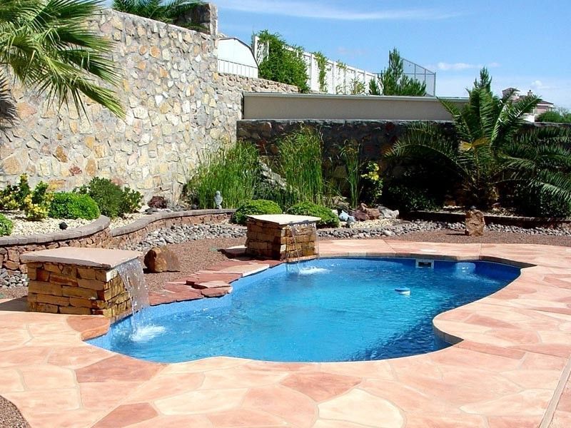 Swimming pool with stone waterfall features and patio, set against a rock wall with greenery.