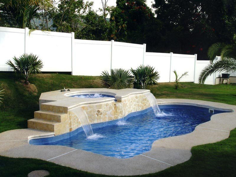 Pool with attached hot tub, waterfall, and stone surround, set in a backyard with white fence and green grass.