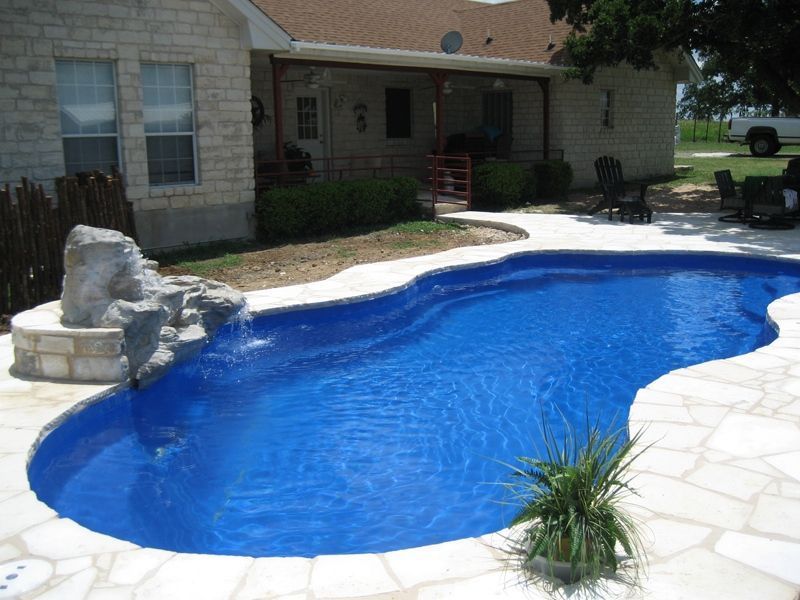 A blue swimming pool with a waterfall, surrounded by light stone, in front of a house.