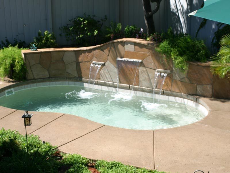 Small, kidney-shaped pool with stone waterfall feature, surrounded by concrete patio and greenery.