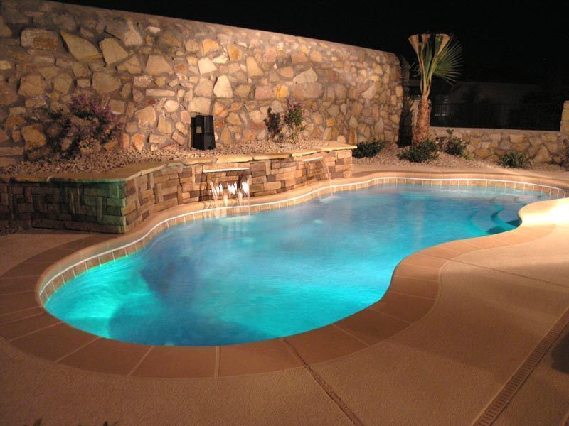 Nighttime view of a turquoise pool with stone wall and a small waterfall feature.