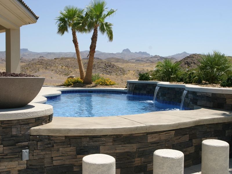 Swimming pool with waterfall feature, stone wall, palm trees, desert mountains in background.