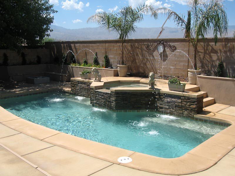 A backyard pool with fountains and tiered waterfall feature, surrounded by tan stone and palm trees.
