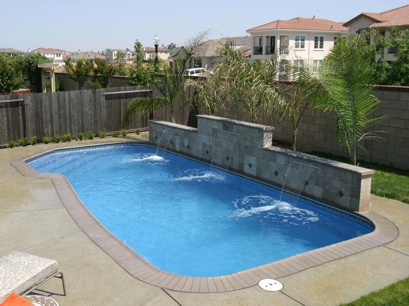 A backyard swimming pool with fountains, surrounded by concrete and a fence; sunny day.