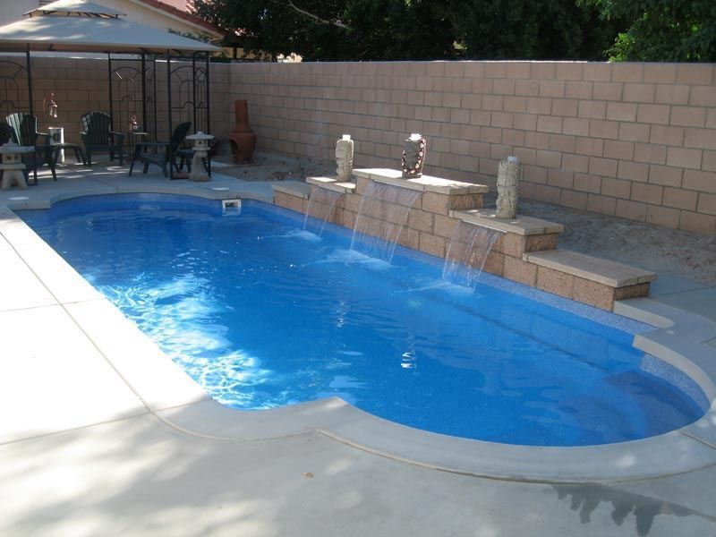 Blue swimming pool with a waterfall feature against a brick wall, patio furniture visible nearby.